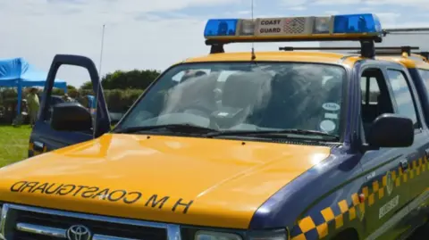 A Coastguard vehicle in yellow and dark blue is branded HM Coastguard on the bonnet, with a blue and white light set-up on the roof. 