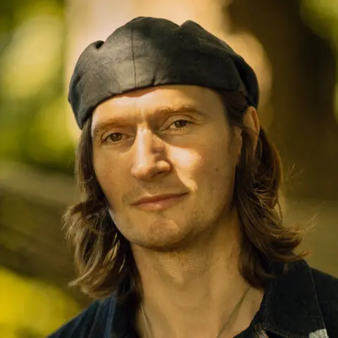 Ross Fraser McLean Erland Cooper has shoulder-length curly brown hair and smiles at the camera in front of a green backdrop. He wears a black hat.