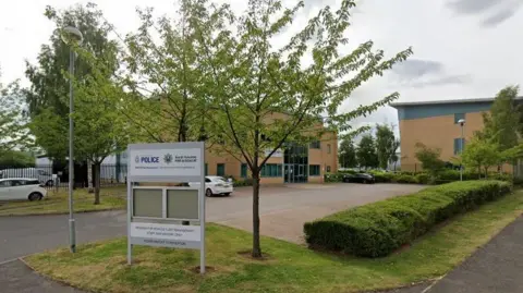 A picture of the building, which is a sandy colour. A car park is situated in front of the building and features several hedges and trees. A sign at the entrance bears the police logo and that of South Yorkshire Fire and Rescue Service.