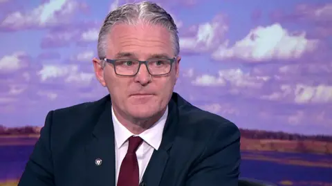 Jarlath Burns - a white/grey haired man with grey glasses on, wearing a navy suit, white shirt and maroon red tie with a GAA pin on his suit lapel