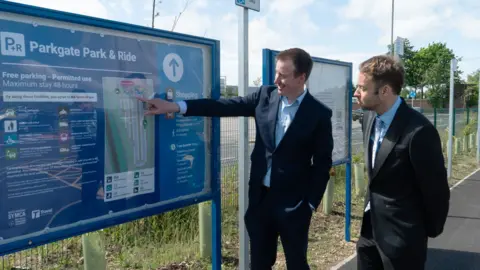 Two men in dark suits are outside, standing next to a large information board headed Parkgate Park & Ride. One of the men is pointing to a map on the board.