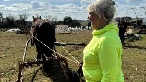 Kate Mobbs-Morgan is standing behind a horse holding its harness. She is wearing a green jumper and is laughing.