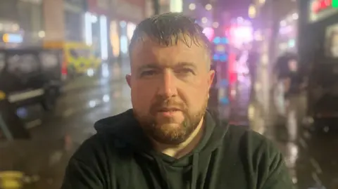 A man stands in the street in the evening. It is wet outside. He is wearing a black hoodie. He has brown and grey hair with a brown short beard. 