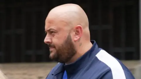 Dominique Saunders is led out of the court. He is wearing a blue tracksuit and is bald with a short dark beard.