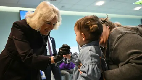 PA Media Queen Camilla interacts with a young child in a hospital setting, reaching out to hold the child's hand as it is supported by its parent. The Queen is wearing a black leather glove