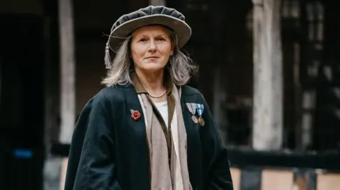 Heidi Meyer wears a large grey cap with a tassel hanging on the right hand side of the cap (the left of the image). She also wears a dark-coloured cloak with a Tudor rose brooch on the right of the cape and her military decorations on the left of her chest.