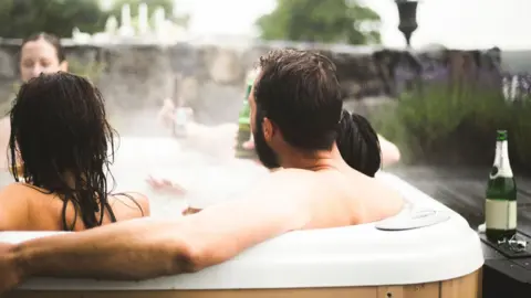Maskot/Getty Images Men and women, seen from the back, relax in an outside hot tub. Green wine bottles can be seen nearby in this stock image.