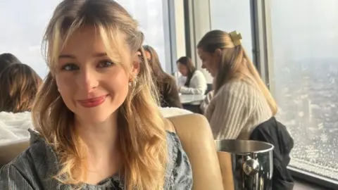 Eve Dulcie sits inside a in a rooftop restaurant with a skyline in the background. She smiles at the camera. She has blonde mid length hair, which is half pulled back from her face with her bangs loose in her face. 
