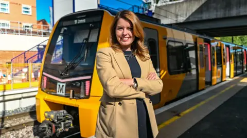 North East Mayor Kim McGuinness standing on a Metro platform with her arms crossed. She is smiling. A yellow and grey Metro train has pulled up at the platform. Its doors are open.