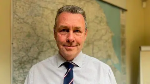 A man is facing the camera, wearing a white shirt and a blue tie. He has short hair and he's stood in his office in front of a map of the Humber region.