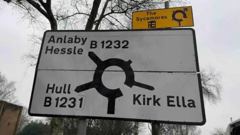 The same road sign has now been cleaned of moss and looks bright white. Bare trees can also be seen under a grey sky.