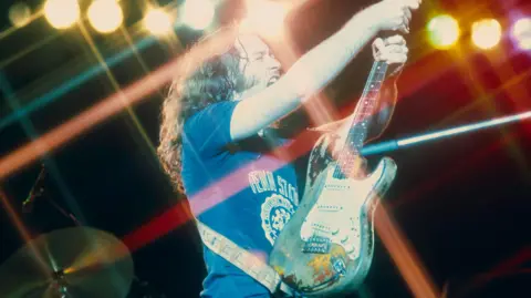 Getty Images Musician Rory Gallagher holds aloft a fender stratocaster guitar, he is wearing a blue t-shirt, and drums can be seen behind him. He has long hair and is standing is in a number of spotlights