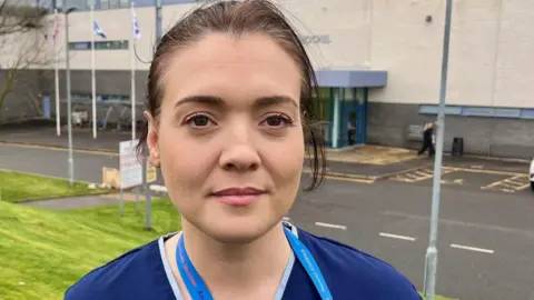 head and shoulders shot of a woman with swept back brown hair. She is wearing a blue uniform and standing in front of a prison building with serious expression on her face.
