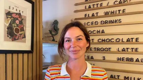 Jenny Morgan wearing a colourful stripy top and stood in front of a board which lists different types of drinks on sale