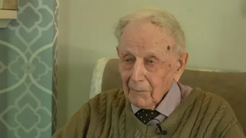 An elderly man with short white hair and wearing a khaki green jumper, a striped shirt and a dark-coloured tie with white spots is sat in an arm chair.
