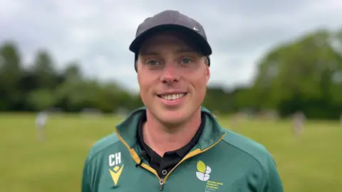 Martin Giles/BBC A man standing outside on a grass pitch and looking directly at the camera and smiling. He is wearing a green zip-up sports jacket and a black cap.
