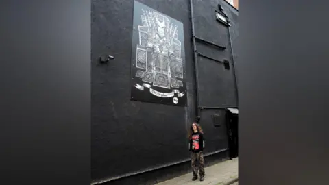 National Album Day A very tall black wall with a mural on the side of a Gryphon in a stylised format of a head with lots of greyish-shaded rectangles surrounding it 