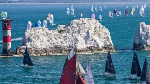 Scores of yachts with their sails up, sailing up to and around the red and white Needles Lighthouse in bright sunshine and blue seas