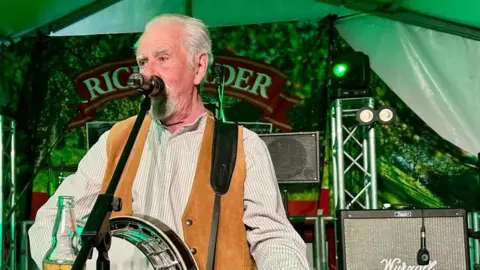 Pete Budd from The Wurzels dressed in a white shirt with brown waistcoat, holding a banjo and singing into a mic. There's stage set and lights behind him inside a green lit marquee