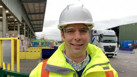 A close-up of councillor Richard Wilkins wearing a high vis jacket and white hard hat, smiling to camera with the recycling centre in the background and a lorry