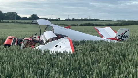 A close-up of the small plane in a field of wheat. It is mostly white livery, with a red strip, and the front of it is completely smashed. On the tail reads: "G-BGRI". In the background are hedges and small pylons, and the sun is starting to set.