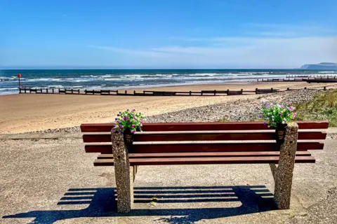 Bettys Mum A bench looking over to the sea at Redcar. The bench has two bunches of colourful flowers tied to it. The ky is blue and cloudless, with the beach stretching into the distance. There are no people in view.