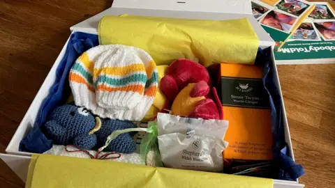 A cardboard box with blue and yellow tissue paper is filled with a knitted bear, a toy dragon, a woolly hat and some biscuits.
 