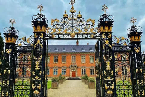Paul Dennis Ornate black and gold wrought iron gates at Tredegar House. The central two gates are open leading through to a gravel walkway with lawns either side and box planters with low bushes in them. The house, a red-brick biulding, is at the end of the walk.