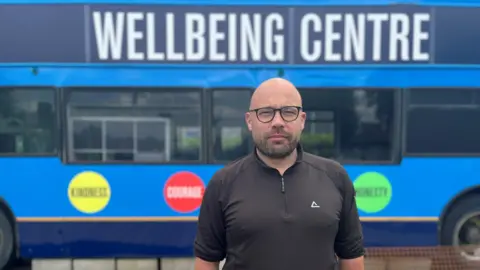 Alex Bish/BBC Matt Paterson standing in front of a blue bus with Welfare Centre in large white text