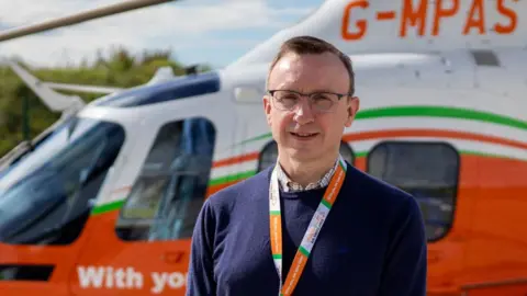 Dr Simon Lewis standing in front of an air ambulance. He has short light brown hair, is wearing glasses and a blue jumper over a checked shirt.