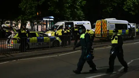 Two police officers walking across the road, with more in the background standing beside a police car and two vans. 