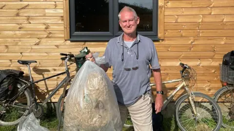 Peter Cunneen Peter Cunneen stands wearing a grey T-shirt with sunglasses on a cord around his neck and pale cargo trousers, holding a big white bin bag containing a destroyed Asian hornet nest. 