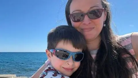 A woman with long brown hair and sunglasses, hugging a young boy who is wearing blue sunglasses. They are standing in front of blue water and a blue sky.