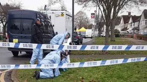 Forensic officers examine the grass behind police tape and by tents, explosives team vans and a police cordon on Willersley Avenue
