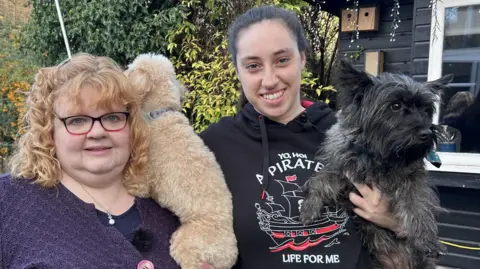 Jane and Victoria stand next to each other in a garden. Jane, on the left, has a navy blue cardigan and top. She has blonde hair and is wearing glasses. Victoria is wearing a black hoodie. They are both smiling. They are both holding dogs. There is a wooden garden shed with a window behind them.