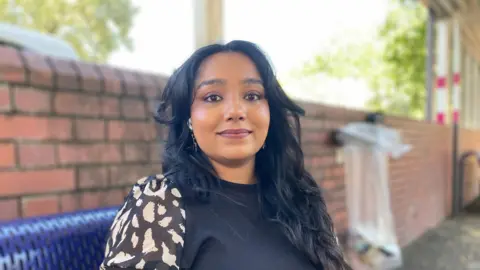 Alex Pope/BBC Feby Thomas sits on a blue metal bench in front of a brick wall. She has dark hair and is wearing a black top over a black and white patterned blouse.