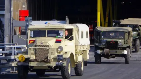 Various military vehicles driving off the ship.