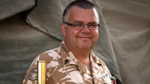 Combat Stress Mark Christian wearing black-rimmed rectangular glasses and a khaki military uniform with a cross on the collar. He is smiling at the camera and there is a green canvas tent behind him.