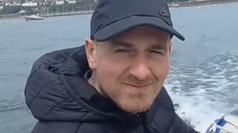 A man in a peaked cap and bomber jacket smiles while steeering a motor boat.