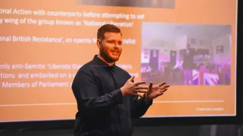 A man wearing a blue shirt is pictured standing in front of an orange screen with writing on it. He is delivering a talk to an audience.