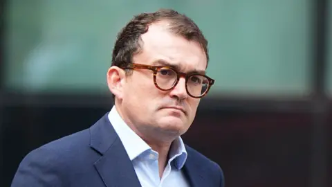 PA Media A man who is looking away from the camera. He has short brown hair, glasses and is wearing a navy blue suit with a light blue shirt.