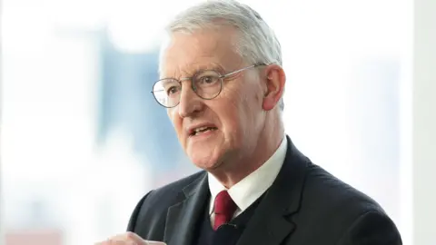 PA Media Hilary Benn wears a black suit, black waistcoat, white shirt and red tie. He wears glasses and has grey hair.