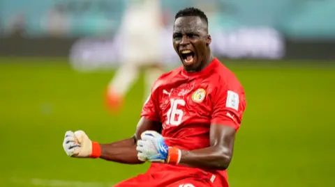 Edouard Mendy in red kit and white gloves screams in celebration while on his knees on a pitch 