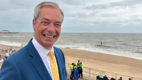 A smiling Nigel Farage wearing a light blue suit, with white shirt and yellow tie. He is stood above a sandy beach 