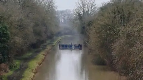 West Midlands Fire Service A canal with a partially-submerged narrowboat positioned across it. A tow path is on the left of the canal with trees either side of the waterway. The canal curves to the right and out of sight in the distance.