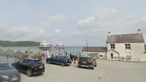 Google images A google street view image shows a beige house on the  waterfront to the right of Garth pier, which has two small circular buildings either side of the start of the pier, which stretches out over the sea towards an greenary covered island in the distance. 