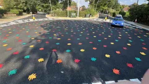 The middle of a roundabout painted with red, orange, green and yellow leaves. A car is entering the circle.