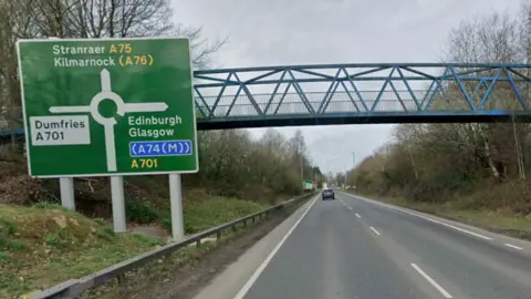 A single-carriageway road with a pedestrian bridge overhead. A sign shows that the road leads up to a roundabout