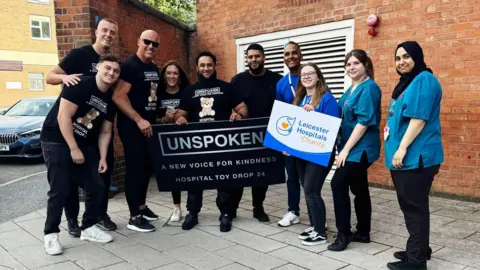 Atiq Rehman Atiq Rehman stands with six of his volunteers, who are dressed in black T-shirts with the Unspoken logo on. They are joined by four other people dressed in blue, and one holding a sign that reads "Leicester Hospitals Charity". 