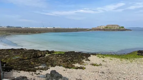 Havelet Bay - a bay with blue sea and in the background is Castle Cornet. In the foreground a pebbly bay with black and brown granite on the left. 